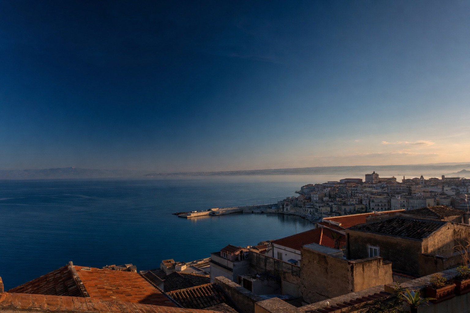 Lo Stretto all'alba visto dall'alto di una cittadina siciliana: acqua calma, tetti in terracotta e cielo blu inchiostro